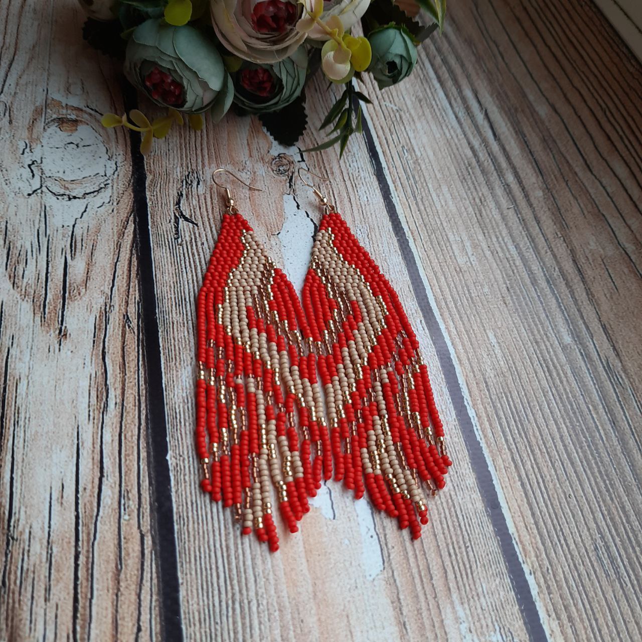 Red and gold beaded earrings on a wooden surface with flowers in the background